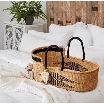 A handwoven tan baby Moses basket or bassinet with a black geometric central pattern and black borders as well as black handles. It is placed on a bed with a teddy leaning on the basket