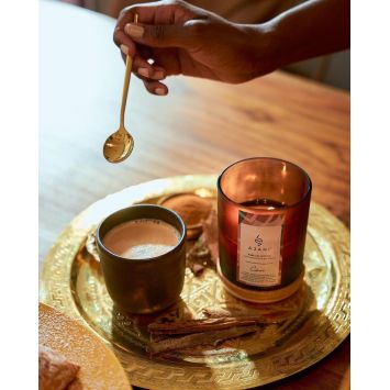 AjanI Chai Scented Candle
next to a black cup of chia on a golden tray with a hand holding a golden spoon above the black cup of chia