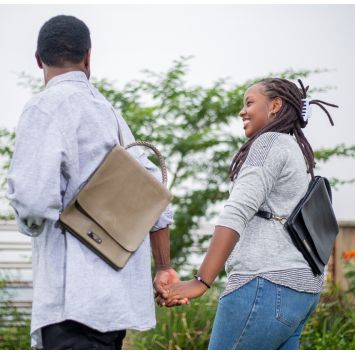 A couple holding hands while leather sling bags are on their backs. One leather sling bag is tan and the other sling bag is black