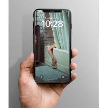 A phone being held out. The lockscreen is an exposed brick background, a bathtub with a teal shower curtain. An arm of someone hanging out of the bathtub. There is a teal bath mat on the floor.